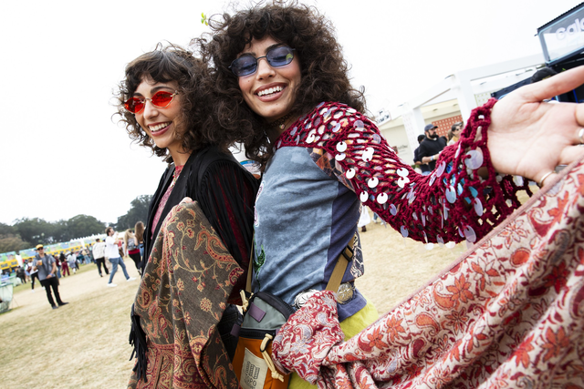 Two women wearing colorful, patterned clothing and sunglasses smile joyfully at an outdoor event, holding hands and embracing.